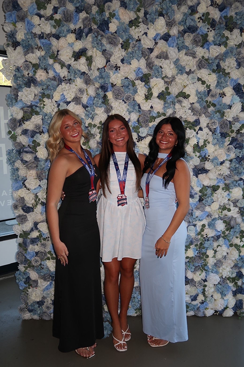 A full-length indoor photo of three young women smiling as they pose together in formal wear against a wall covered in white, light blue, and grey artificial roses. The woman on the left wears a strapless black floor-length dress, the center woman wears a sleeveless white mini dress, and the woman on the right wears a sleeveless light blue maxi dress. All three wear competition lanyard medals around their necks.