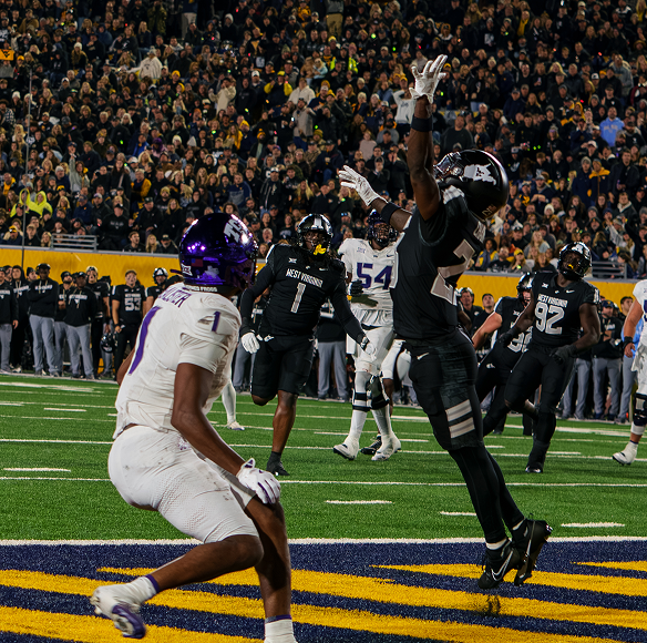A night college football action scene at a packed stadium. A West Virginia player in a black uniform leaps high with arms extended in the end zone to defend a pass, while a TCU receiver in a white and purple uniform (#4) looks on from below. Other players and a packed crowd filling the stands are visible in the background.