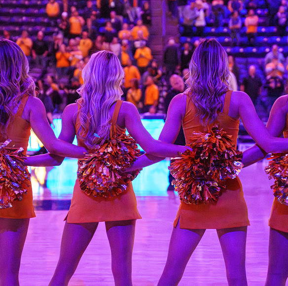 A back-view photograph of four cheerleaders standing in a line on a basketball court under vibrant purple lighting. They are dressed in matching orange sleeveless dresses and hold shiny metallic pom-poms behind their lower backs with arms linked together. A crowded arena seating section is visible blurred in the background.