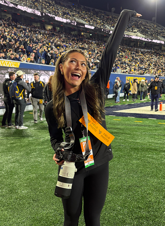 A candid full-length photo of photographer Kylie Yobe standing on the sidelines of a football field at night. She smiles exuberantly with one arm raised in celebration, wearing a black long-sleeve top, dark leggings, and media credentials—including an orange FIELD PHOTO pass—around her neck. She holds a professional camera equipped with a large white telephoto lens, with a packed, lit stadium crowd filling the background.
