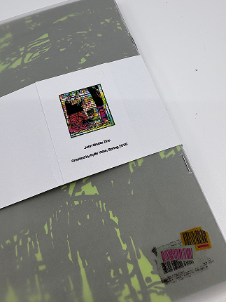 A high-angle photograph of a physical zine booklet with a gray and lime-green patterned cover. The zine is wrapped with a white paper belly band featuring a square, colorful stained-glass style illustration centered above the text John Waters Zine and Created by Kylle Yabe, Spring 2026. A small barcode label sticker is visible in the bottom-right corner.