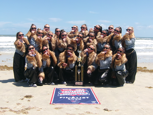 Outdoor group portrait of a dance team posing on a sunny sandy beach with the ocean waves in the background. The team members are wearing matching fur-trimmed jackets, dark pants, and sunglasses, proudly displaying their medals around their necks or in hand. In the foreground, a large championship trophy stands in the center above an NDA/NCA Collegiate Cheer and Dance Championship finalist banner laid flat on the sand.