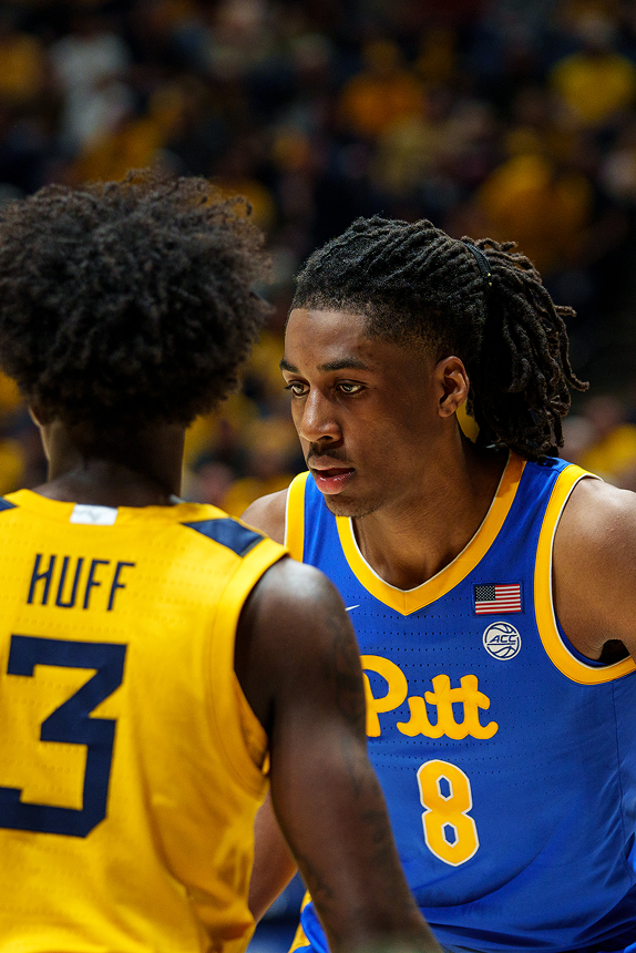 Vertical sports action photograph capturing a basketball player in a royal blue and gold uniform executing a jump shot with a high follow-through. A West Virginia player in a bright gold uniform leaps with an outstretched arm attempting to block the shot as the orange basketball floats at the apex of its trajectory. A packed arena crowd fills the background blurred in the distance.