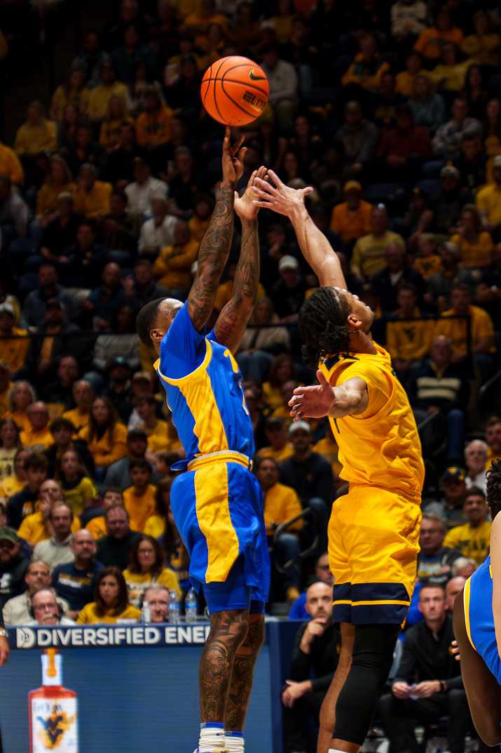 A candid post-game sports photograph capturing a West Virginia men's basketball player wearing number 23 in a bright gold uniform. He smiles widely and reaches out to high-five fans, highlighted by dramatic, vibrant purple and pink stadium lighting that casts dynamic rim light across his face and shoulder against a blurred arena background.