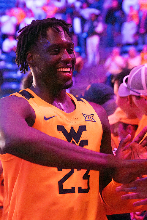 A full-length court-side action photograph featuring the West Virginia Mountaineer mascot in buckskins and coonskin cap, standing on the basketball court with arms raised high, holding his musket rifle in celebration. He is flanked by WVU cheerleaders holding gold pom-poms, set against the backdrop of the basketball hoop, shot clock, and a packed arena crowd dressed in gold and navy.