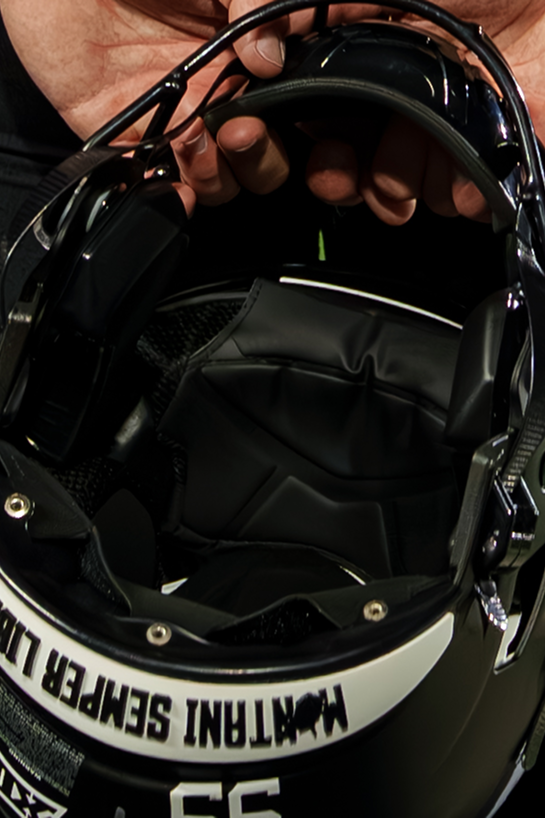 Detailed close-up photo looking inside a black football helmet held by a pair of hands at the top. The image highlights the dark interior padding, ear pads, and facemask structure. Along the rear bumper of the helmet, white lettering displays the West Virginia state motto MONTANI SEMPER LIBERI alongside a football graphic and the number 55 on the lower shell.