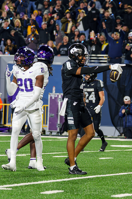 On-field action photograph during a college football game. On the right, a West Virginia player in a black uniform and helmet points dramatically forward with one hand while holding a football in the other to signal a first down. On the left, TCU defensive players wearing white jerseys with purple numbers and purple helmets stand nearby. A cheering crowd in the stadium stands fills the background.