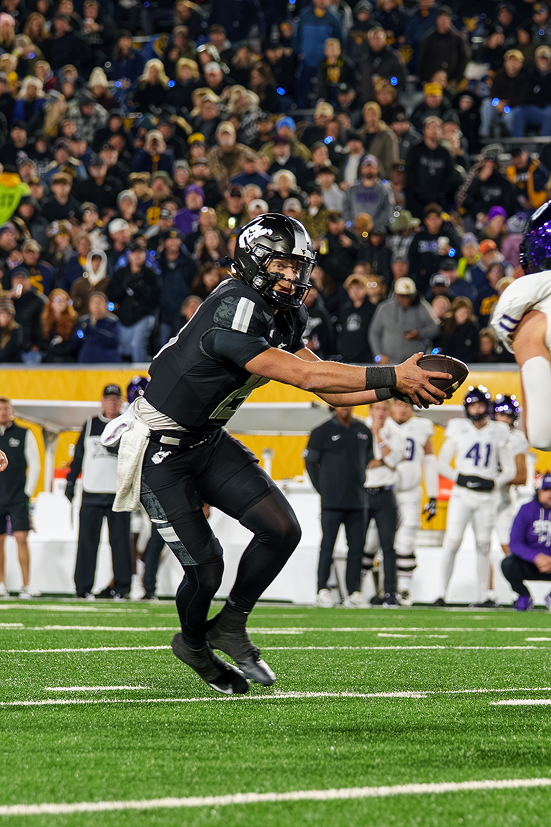 Full-length action photograph of a West Virginia player in a black uniform and black helmet leaning forward low to execute a handoff with both hands on the football. The game takes place on green turf at night, with sideline personnel and a blurred crowd filling the background stadium stands.