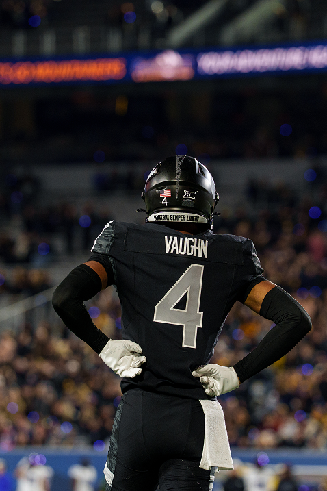Rear-view photograph of West Virginia football player number 4, VAUGHN,wearing a black jersey, black arm sleeves, white gloves, and a black helmet. He stands with his hands on his hips looking out toward the field, with the blurred lights and crowds of a stadium setting in the background.