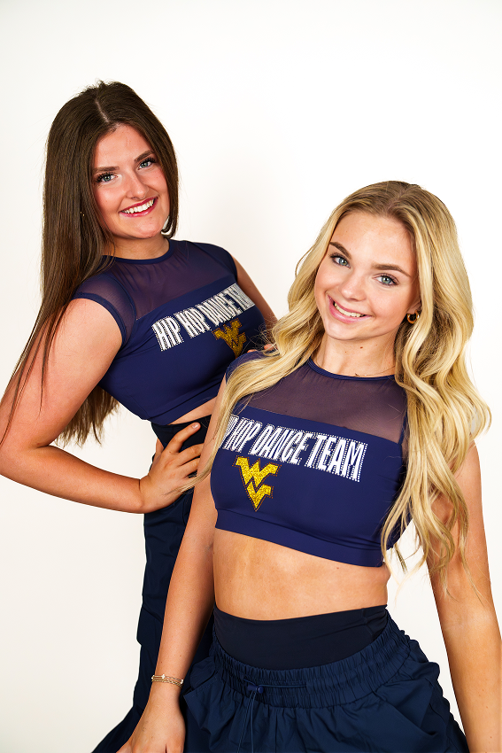 Studio photo of two dancers posing against a seamless white backdrop. Both are wearing matching navy crop tops featuring mesh necklines, a gold WV logo, and HIP HOP DANCE TEAM in glittery white letters, paired with navy pants. A brunette dancer stands in the back with her hand on her hip, while a blonde dancer stands slightly in front, both smiling directly at the camera.
