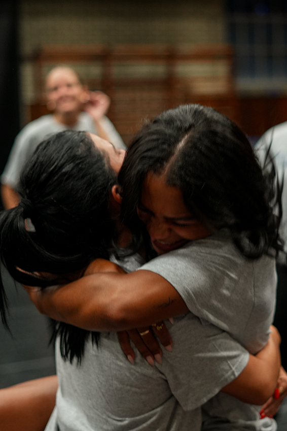 A close-up candid photograph capturing an emotional hug between two team members wearing light gray t-shirts. One dancer with dark wavy hair embraces her teammate tightly with a joyous, tearful smile, arms wrapped around each other in a dance studio setting with blurred figures in the background.